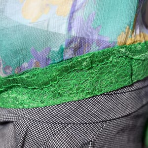 Op de afbeelding: Close-up van een kleurrijk kledingstuk met een bloemenprint in geel, paars en teal. Een heldergroene kanten rand scheidt de stof met patroon van een zwart-wit geruite stof.