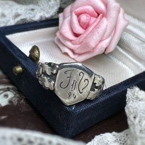 Peut inclure: Bague en argent avec une face en forme de bouclier, gravée des initiales "F2" et du chiffre "84". La bague est présentée dans un écrin bleu foncé, avec une rose rose en arrière-plan. La bague a une esthétique vintage et antique.