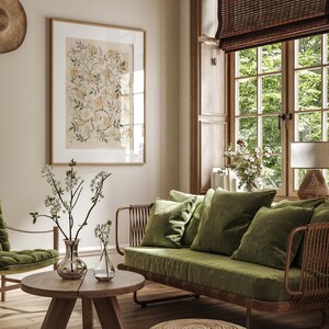 May include: A green velvet sofa with a woven rattan frame sits in a living room with a wooden floor. The sofa has three pillows and is positioned in front of a window with a bamboo blind. A small round wooden coffee table sits in front of the sofa with two clear glass vases filled with white flowers. A small woven ottoman sits in front of the sofa.
