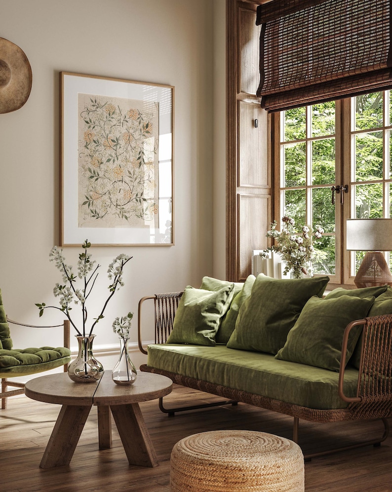 May include: A green velvet sofa with a woven rattan frame sits in a living room with a wooden floor. The sofa has three pillows and is positioned in front of a window with a bamboo blind. A small round wooden coffee table sits in front of the sofa with two clear glass vases filled with white flowers. A small woven ottoman sits in front of the sofa.