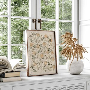 May include: A framed botanical print with a floral design in shades of green, white, and yellow. The print is displayed on a white shelf with a vase of dried grasses and a stack of books.
