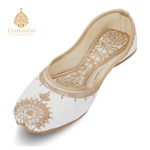 May include: A white flat shoe featuring intricate gold embroidery on the toe and along the collar. The design includes circular floral patterns and decorative stitching. The interior is lined in a light beige material with a matching gold trim.