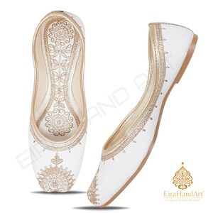 May include: A pair of white flat shoes featuring intricate gold embroidery on the toe and along the inner sole. The design includes detailed floral and circular patterns in metallic gold thread. The shoes have a pointed toe and a slim profile.