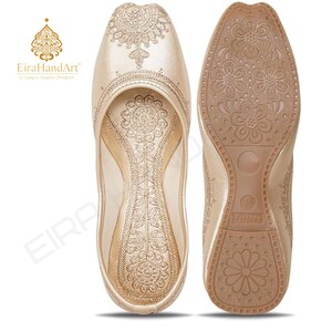 May include: A pair of champagne-coloured slip-on flats featuring intricate gold embroidery on the upper and interior sole. The outsole is tan with a repeating floral pattern and a visible size 10 marking. The EiraHandArt logo is present.