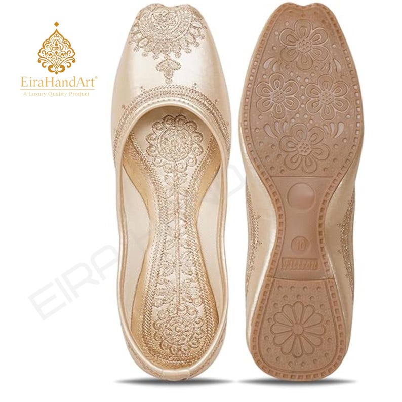 May include: A pair of champagne-coloured slip-on flats featuring intricate gold embroidery on the upper and interior sole. The outsole is tan with a repeating floral pattern and a visible size 10 marking. The EiraHandArt logo is present.