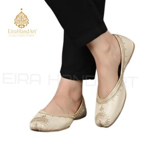 May include: A pair of cream-coloured slip-on flats featuring intricate gold embroidery on the rounded toe and along the side seams. The footwear has a closed-toe design with a low profile and a smooth finish, accented by subtle gold stitching details.