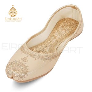 May include: A beige flat shoe featuring intricate gold embroidery on the toe and along the inner sole. The design includes detailed floral patterns stitched with metallic thread, a rounded toe, and a low-cut silhouette with a smooth finish.