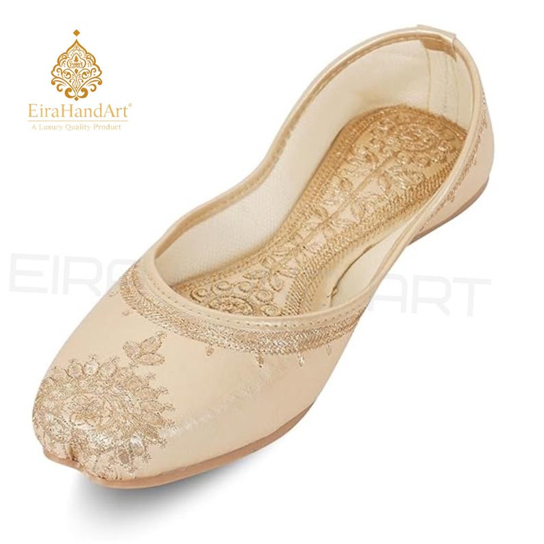 May include: A beige flat shoe featuring intricate gold embroidery on the toe and along the inner sole. The design includes detailed floral patterns stitched with metallic thread, a rounded toe, and a low-cut silhouette with a smooth finish.