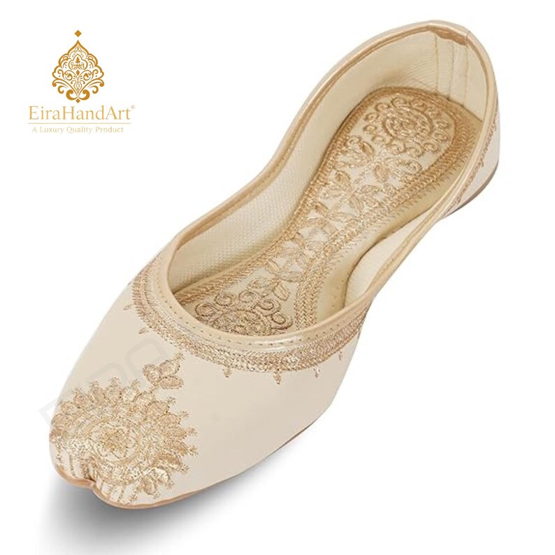 May include: A cream-coloured flat shoe featuring intricate gold thread embroidery on the toe and inner sole. The design includes floral-inspired patterns and a rounded toe shape. The brand name EiraHandArt is visible in the upper left corner.