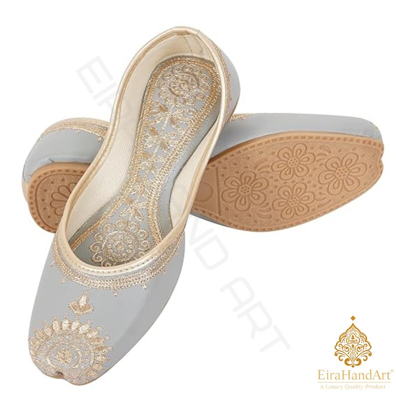 May include: A pair of light grey flat shoes featuring intricate gold embroidery on the toe and along the inner sole. The shoes have a rounded toe design and a tan-coloured sole with a repeating floral pattern embossed on the bottom surface.