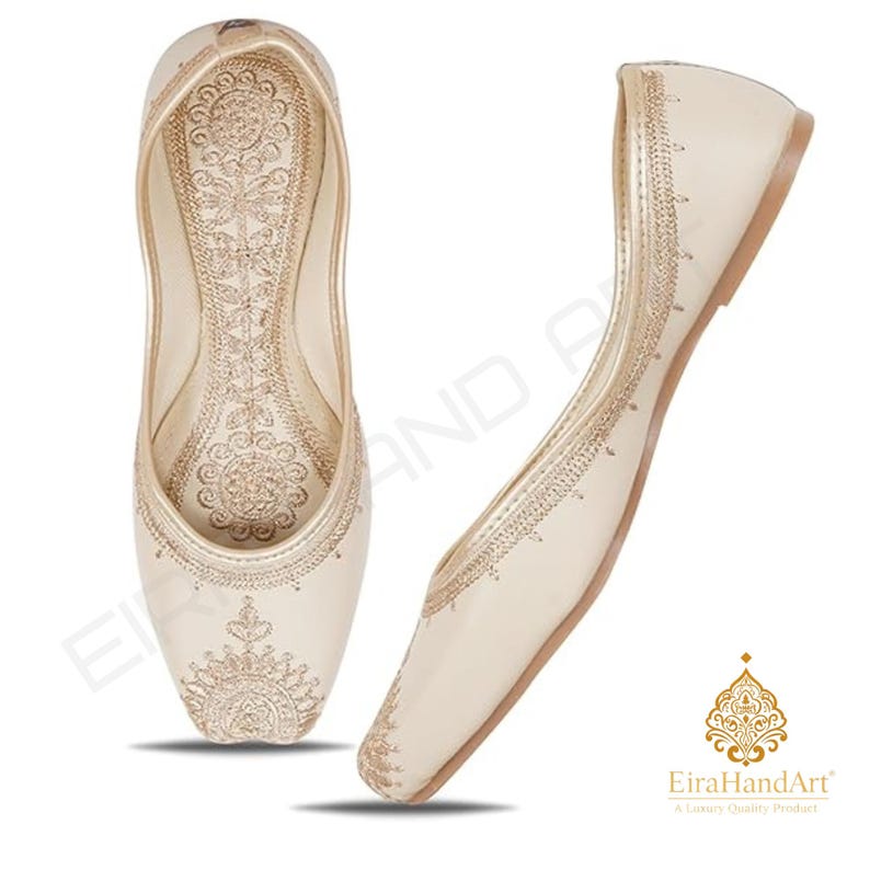 May include: A pair of cream-coloured flat shoes featuring intricate gold embroidery on the toe and along the side edges. The interior sole displays a detailed floral pattern, and the EiraHandArt logo is visible in the bottom right corner.
