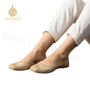 May include: A pair of beige slip-on flats featuring intricate gold embroidery on the rounded toe area. The shoes have a low-cut silhouette and a smooth finish, paired with white cropped trousers. The EiraHandArt logo is visible in the corner.