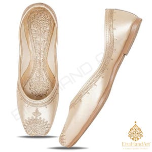 May include: A pair of champagne-coloured flat shoes featuring intricate gold-toned embroidery on the toe and inner sole. The design includes a pointed toe and a low-profile silhouette, with a side view showing the smooth exterior and stitched detailing.