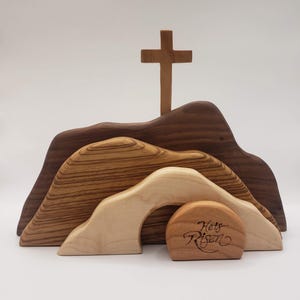 May include: Wooden Easter tomb with a cross on top. The tomb is open and the stone is rolled away. The stone has the words "He's Risen" engraved on it.