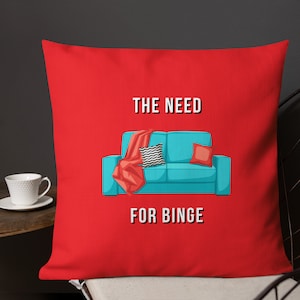 May include: Red throw pillow with the text "THE NEED FOR BINGE". The pillow features a teal couch graphic with a red blanket, a black and white patterned pillow, and a red pillow.