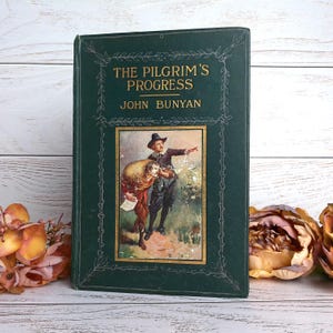 May include: A vintage hardcover book titled "The Pilgrim's Progress" by John Bunyan. The cover is dark green with gold lettering and a framed illustration of two men. The book is set against a white wooden surface with floral accents.
