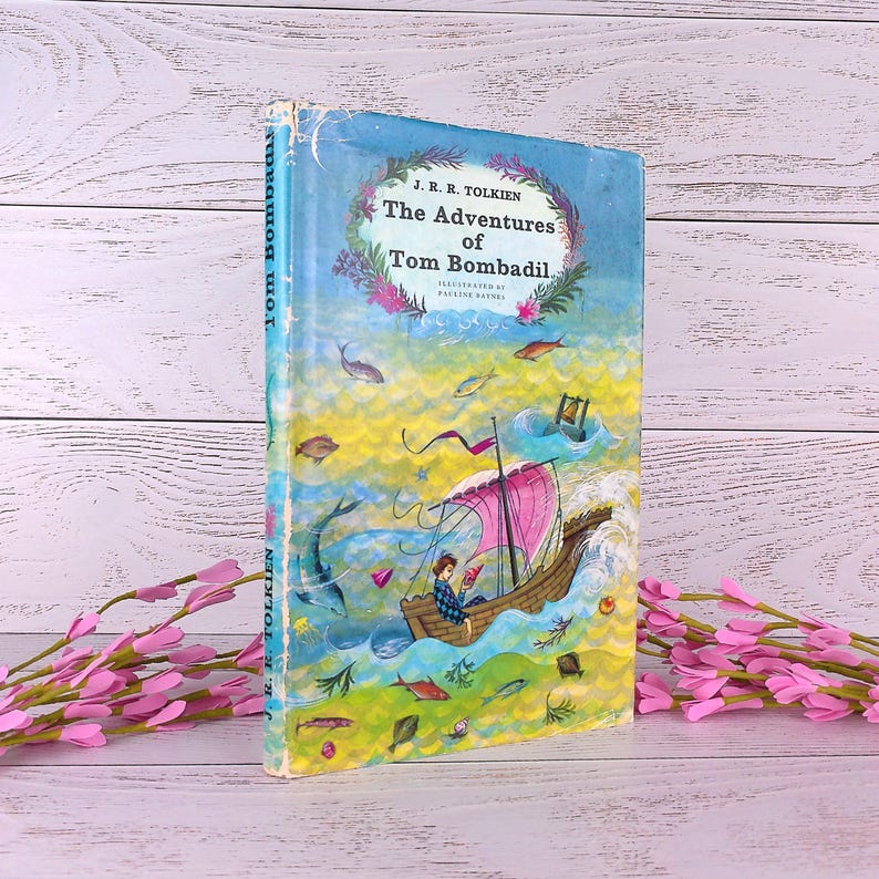 Op de afbeelding: Vintage hardcover boek getiteld "The Adventures of Tom Bombadil" van J.R.R. Tolkien. De omslag toont een kleurrijke illustratie van een boot die op golvend water vaart, met vissen en een lezende persoon. De titel van het boek staat in een bloemenkrans.