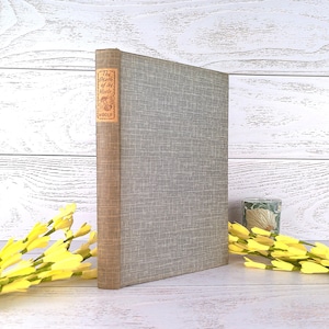 Puede incluir: Un libro de tapa dura titulado "The Death of the Moth" de Woolf. El libro tiene una cubierta gris claro con un patrón texturizado y un lomo marrón. El título está en una etiqueta roja y marrón. Flores amarillas en primer plano.
