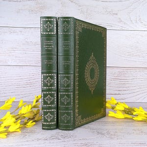 Pode incluir: Dois livros de capa dura verde-escuro intitulados Charles Dickens Complete Works Christmas Stories I e II. Os livros apresentam padrões de filigrana dourada nas lombadas e na capa, com nervuras e motivos florais decorativos nas lombadas.
