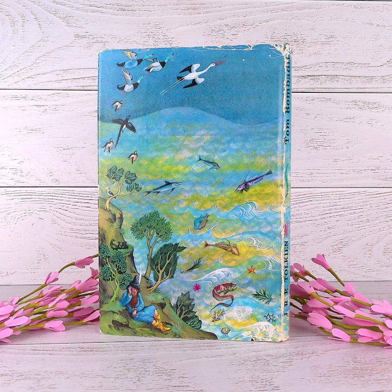 Op de afbeelding: Een vintage boek met een kleurrijke illustratie. De omslag toont een tovenaar, vogels en zeedieren tegen een blauwgroene achtergrond. De titel "Tom Bombadil" en de auteur "J.R.R. Tolkien" zijn zichtbaar op de rug.