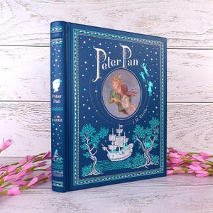 May include: A dark blue hardcover book titled "Peter Pan" with illustrations of Peter Pan, a ship, and fairies. The spine displays the title and author, J.M. Barrie. The book is set against a white wooden background. The book is a classic children's story.