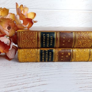 Könnte beinhalten: Zwei antike Bücher mit goldgeprägten Einbänden und schwarzen Rückseiten. Die Bücher tragen den Titel "Cowper's Poetical Works" und sind die Bände I und II. Die Bücher liegen auf einer weißen Holzoberfläche mit einem Strauß orangefarbener Blumen.