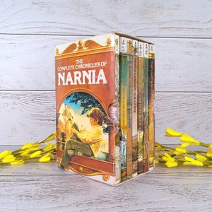 May include: A boxed set of The Complete Chronicles of Narnia books. The box is orange and brown with the title in large white letters. The book covers are visible, each with unique illustrations and titles. Yellow flowers are in the foreground.