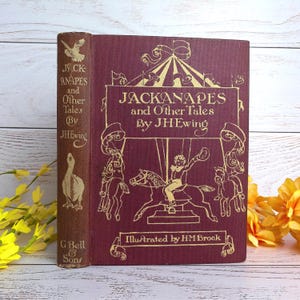 Op de afbeelding: Een vintage hardcover boek getiteld "Jackanapes and Other Tales" van J.H. Ewing, geïllustreerd door H.M. Brock. De omslag is bordeauxrood met gouden letters en illustraties van een carrousel en een gans. De rug is bruin.