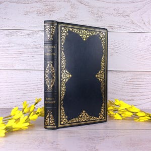 Puede incluir: Un libro de tapa dura negro titulado Wuthering Heights de Emily Bronte, con intrincados patrones de filigrana dorada en la portada y el lomo. El lomo muestra el título y el nombre de la autora en letras doradas sobre un fondo oscuro.