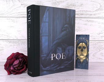Può includere: Un libro con copertina rigida intitolato "POE TALES & POEMS" con un'illustrazione scura di un corvo. È presente anche un segnalibro decorativo con un disegno di scarabeo dorato. Una rosa rosso scuro è a sinistra.