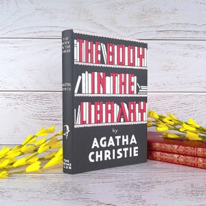 Könnte beinhalten: Ein Buch mit dem Titel "The Body in the Library" von Agatha Christie. Das Buch hat einen schwarzen Einband mit weißem und rotem Text und einem Regaldesign. Gelbe Blumen sind im Vordergrund.