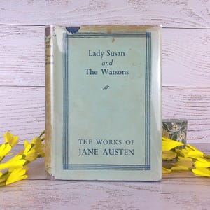 May include: A vintage book titled "Lady Susan and The Watsons" by Jane Austen. The book has a light blue cover with dark blue border and text. The book is standing upright on a light wood surface, with yellow flowers in the foreground.
