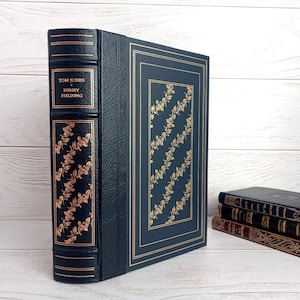 Puede incluir: Un libro de tapa dura de color azul oscuro con letras doradas en el lomo. El título es "Tom Jones" de "Henry Fielding". El libro está sobre una superficie de madera blanca.