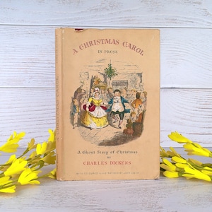 Może przedstawiać: Zabytkowa książka w twardej oprawie zatytułowana A Christmas Carol in Prose, A Ghost Story of Christmas autorstwa Charlesa Dickensa. Beżowa okładka zawiera centralną kolorową ilustrację przedstawiającą ludzi tańczących na przyjęciu oraz czerwony tekst.