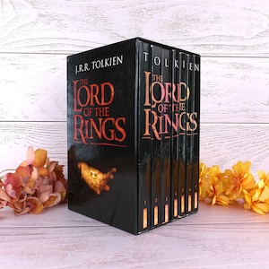 Puede incluir: Un estuche de libros "El Señor de los Anillos" de J.R.R. Tolkien. El estuche negro presenta el título en letras naranjas. Los lomos de los libros son visibles, cada volumen está numerado. Flores decorativas en primer plano.