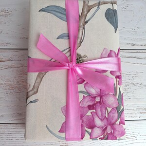 Op de afbeelding: Een rechthoekig cadeau verpakt in stof met bloemmotieven, roze orchidee&euml;n, grijsgroene bladeren en bruine takken. Het cadeau is vastgebonden met een felroze satijnen lint, dat een strik vormt. De achtergrond is een wit houten oppervlak.