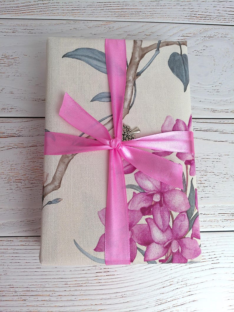 Op de afbeelding: Een rechthoekig cadeau verpakt in stof met bloemmotieven, roze orchidee&euml;n, grijsgroene bladeren en bruine takken. Het cadeau is vastgebonden met een felroze satijnen lint, dat een strik vormt. De achtergrond is een wit houten oppervlak.