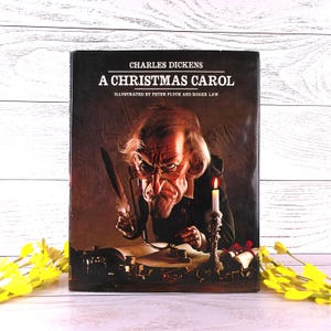 Pode incluir: Um livro de capa dura intitulado A Christmas Carol de Charles Dickens, ilustrado por Peter Fluck e Roger Law. A capa apresenta uma caricatura de um homem com uma pena numa secretária com uma vela acesa, tinteiros e pergaminhos.
