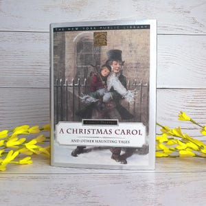 Pode incluir: Um livro de capa dura intitulado A Christmas Carol and Other Haunting Tales de Charles Dickens, com uma etiqueta Collector's Edition da New York Public Library. A capa mostra uma pessoa de cartola carregando uma criança em um cenário nevado.