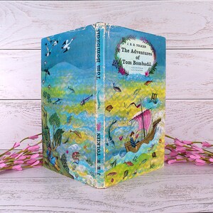 Op de afbeelding: Een vintage hardcover boek getiteld "The Adventures of Tom Bombadil" van J.R.R. Tolkien. De omslag toont een grillige illustratie met een boot, vissen en een kleurrijk landschap in tinten blauw, groen en geel.