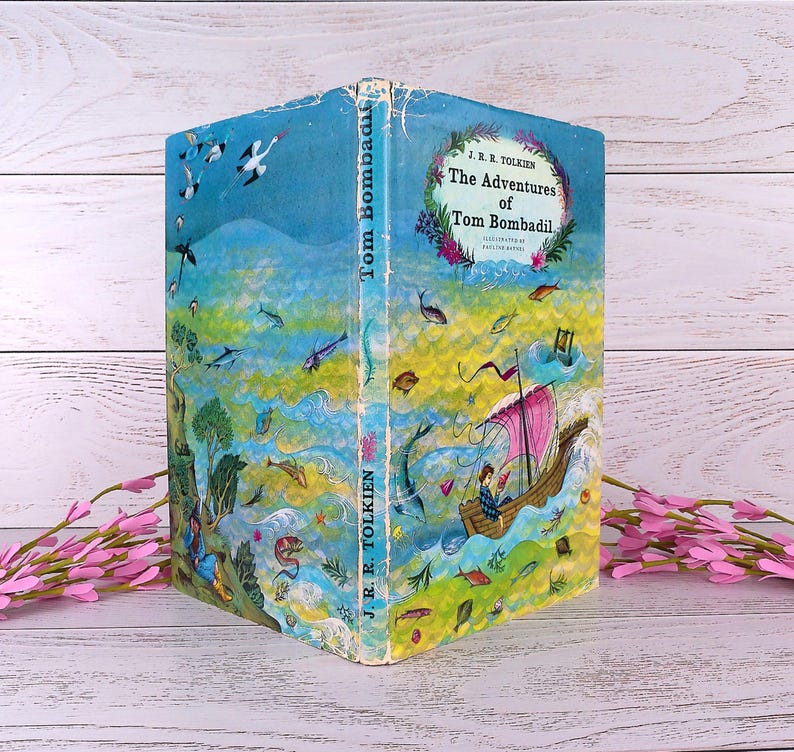 Op de afbeelding: Een vintage hardcover boek getiteld "The Adventures of Tom Bombadil" van J.R.R. Tolkien. De omslag toont een grillige illustratie met een boot, vissen en een kleurrijk landschap in tinten blauw, groen en geel.