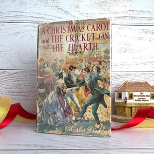 Pode incluir: Livro de capa dura vintage intitulado "A Christmas Carol and The Cricket on the Hearth" de Charles Dickens. A capa apresenta uma ilustração colorida de uma cena de baile com pessoas dançando. O livro tem uma aparência desgastada.