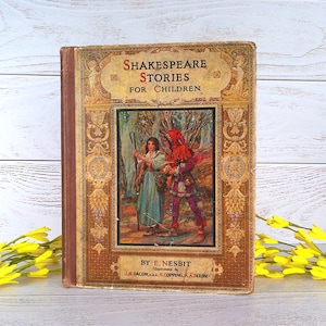 Puede incluir: Libro antiguo, "Shakespeare Stories for Children" de E. Nesbit. La portada presenta una ilustración de dos figuras en un bosque. El libro tiene una lomo marrón y un borde decorativo.