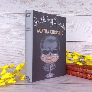 Könnte beinhalten: Ein gebundenes Buch mit dem Titel "Sparkling Cyanide" von Agatha Christie. Das Buch hat einen schwarzen Einband mit einem Totenkopf und einem Cocktailglas. Der Buchrücken ist hellgrau. Gelbe Blumen und weitere Bücher sind im Hintergrund.