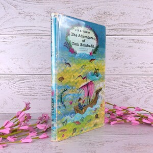 Op de afbeelding: Een hardcover boek getiteld "The Adventures of Tom Bombadil" van J.R.R. Tolkien. De omslag toont een kleurrijke illustratie van een boot op het water, omringd door vissen en een bloemenrand. Het boek is verpakt in doorzichtig plastic.