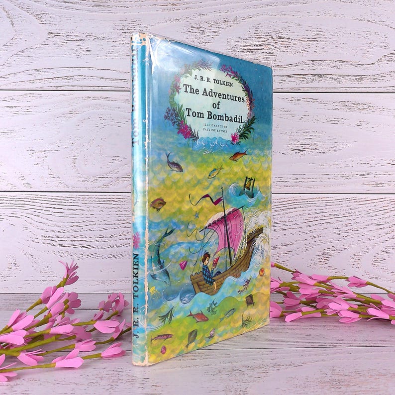 Op de afbeelding: Een hardcover boek getiteld "The Adventures of Tom Bombadil" van J.R.R. Tolkien. De omslag toont een kleurrijke illustratie van een boot op het water, omringd door vissen en een bloemenrand. Het boek is verpakt in doorzichtig plastic.