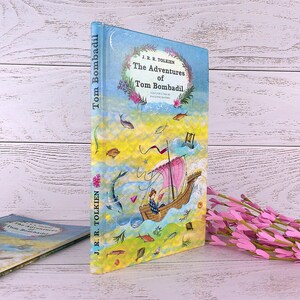 Op de afbeelding: Een hardcover boek getiteld "The Adventures of Tom Bombadil" van J.R.R. Tolkien. De omslag toont een kleurrijke illustratie van een boot met een roze zeil, vissen en een lezende persoon. Het boek staat rechtop naast een kleiner, vergelijkbaar boek.