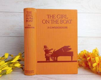 May include: An orange hardcover book titled "The Girl on the Boat" by P.G. Wodehouse. The book cover features a silhouette of a woman playing a piano.