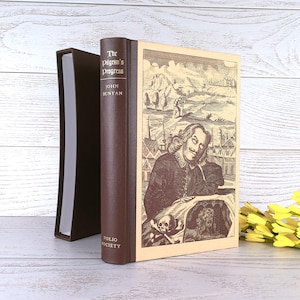 May include: A brown and cream hardback book titled The Pilgrim's Progress by John Bunyan, published by Folio Society. The cover features a detailed black and white illustration of a person, a skull, and a landscape. It includes a matching brown slipcase.