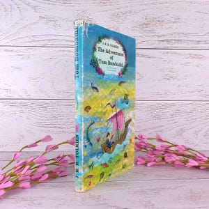 Op de afbeelding: Een vintage exemplaar van "The Adventures of Tom Bombadil" van J.R.R. Tolkien. De boekomslag toont een kleurrijke illustratie van een boot die op het water vaart, met de titel in een bloemenkrans. De rug van het boek luidt "Tom Bombadil" en "J.R.R. Tolkien."
